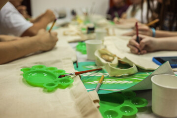 Close-up of art workshop with paintbrushes, green plastic palettes, and people painting on fabric bags at a shared table, creative group activity