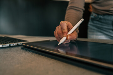 Close-up of hand holding stylus and working on digital tablet with laptop in background on desk