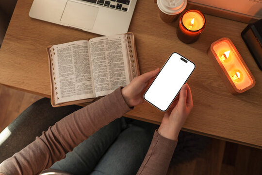 Person holding a smartphone with a blank white screen at a wooden desk, next to an open Bible, a laptop, a coffee cup, and lit candles, creating a warm and peaceful study or devotional setting