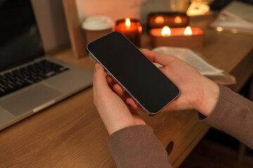 Close-up of hands holding a smartphone with a black screen at a wooden desk, next to a laptop, lit candles, a takeaway coffee cup, and an open book in the background