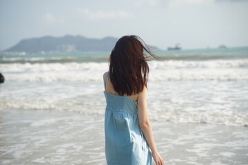 A woman in a light blue dress stands on a beach, facing the sea. Her long hair blows in the wind, with waves and distant islands in the background