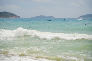 一幅海滩场景，海浪拍打着海岸。大海呈现出蓝色和绿色的色调，在明亮的天空下，远处有船只和人群。