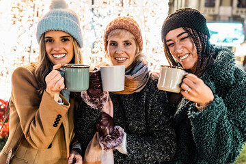 Three diverse friends looking at camera with mugs at a Christmas market. Happy multicultural group toasting with hot chocolate and enjoying the festive holiday season at night.