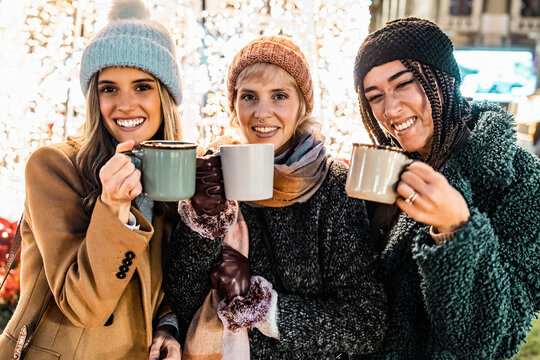 Three diverse friends looking at camera with mugs at a Christmas market. Happy multicultural group toasting with hot chocolate and enjoying the festive holiday season at night.
