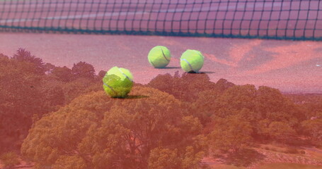 Fototapeta premium Resting three yellow tennis balls on red clay court, with tennis net and forest overlay