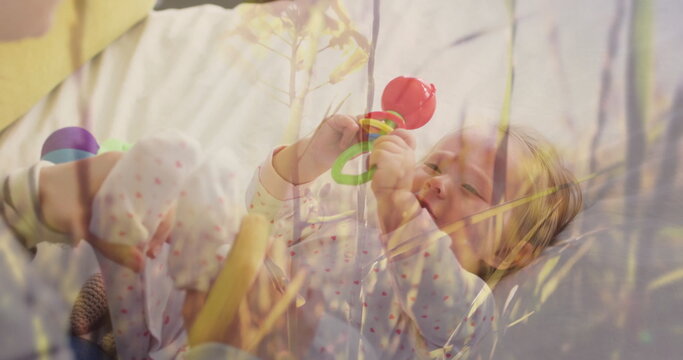 Gazing infant girl in pajamas gripping red and green rattle on bed in nursery, plant reflection