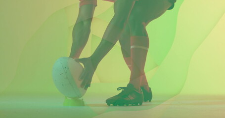 Rugby player's legs steadying white rugby ball on green tee in studio, with green-yellow backdrop