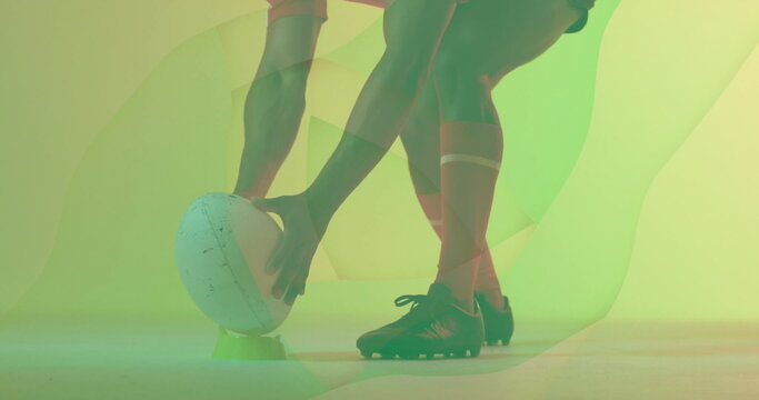 Rugby player's legs steadying white rugby ball on green tee in studio, with green-yellow backdrop - Powered by Adobe