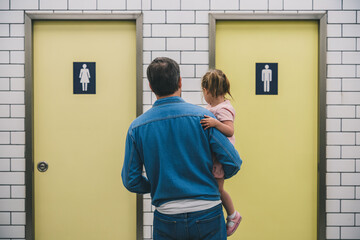Fototapeta premium Father with young daughter hesitating in front of men’s and women’s restroom doors. Concept of gendered public toilet challenges for parents