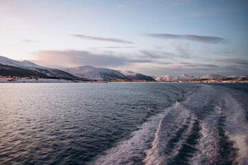 The powerful wake of a boat cuts through a dark sea, leading the eye towards a distant, panoramic view of a snowy mountain range and coastal town lights at dusk.
