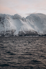 A powerful, close-up view of a vast, snow-covered mountain rising directly from the dark, choppy water of a fjord, with a soft, minimalist sky above.