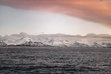 A panoramic landscape featuring a vast, dark sea under a dramatic sky with a large, sweeping cloud formation and a distant range of snow-capped mountains.