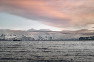 A panoramic landscape featuring a vast, dark sea under a dramatic sky with a large, sweeping cloud formation and a distant range of snow-capped mountains.