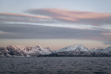 A breathtaking panoramic shot of a snow-covered mountain range reflected in a dark, choppy sea, all bathed in the warm, pastel colors of a stunning sunset.
