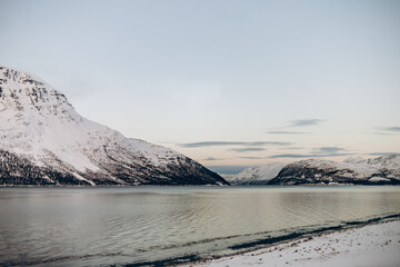 A serene winter landscape with a calm fjord framed by steep, snow-covered mountains under a soft, twilight sky with subtle clouds.