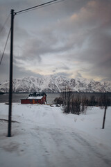 A secluded red hut on a snowy shoreline, with a serene fjord and a magnificent range of snow-covered mountains stretching across the horizon under a soft, moody sky.
