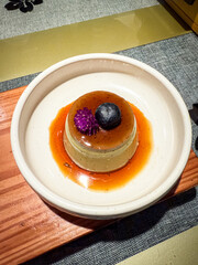 Japanese caramel pudding topped with a blueberry and purple edible flower, served on a ceramic plate with rich caramel sauce.