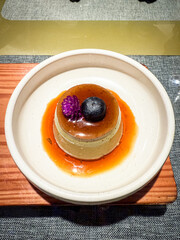 Japanese caramel pudding topped with a blueberry and purple edible flower, served on a ceramic plate with rich caramel sauce.