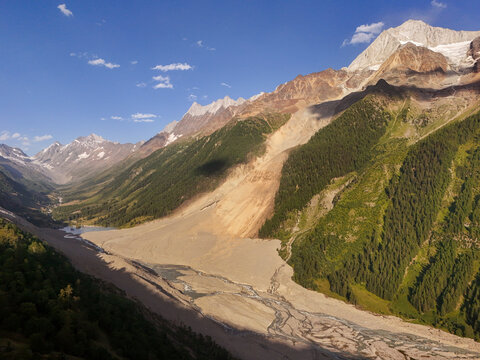 Aerial view of a valley floor carved by glacial streams, flanked by verdant forests and rising to snow-capped peaks under a clear sky, Blatten, Wallis, Switzerland.
