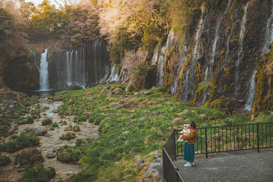 Happy Asian mother holding excited little daughter enjoying trip near Shiraito waterfall in Japan during spring season – cheerful family travel, joyful childhood, nature experience and bonding moment.
