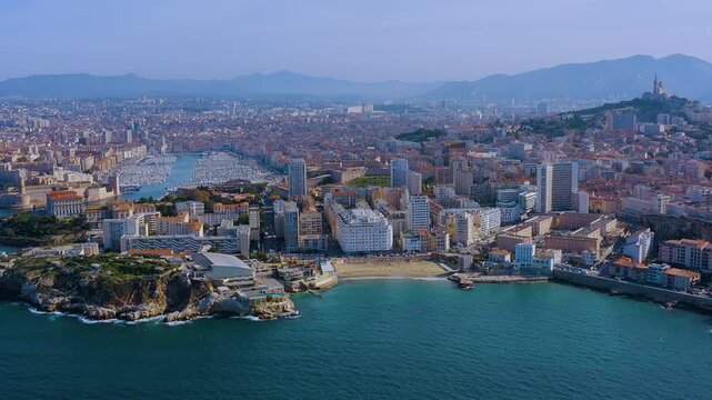 4K drone panorama of Marseille: Vieux-Port packed with yachts, Catalans Beach and Palais du Pharo at the entrance, Notre-Dame de la Garde on the hill, Mediterranean coast and Provence hills beyond.