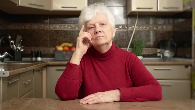 Elderly woman places finger on temple. Old female sits thoughtfully in kitchen. Senior woman is deep in thought, immersed in reflections while alone at home. Flabby and wrinkled skin on senior female