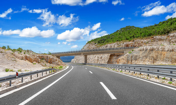 A modern concrete bridge spans over a curved highway surrounded by rocky hills under a bright blue sky with scattered clouds.
