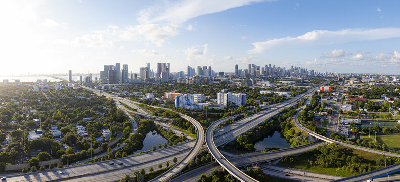 Aerial view of a complex highway interchange slicing through lush green spaces towards the gleaming skyline of downtown Miami, Florida, United States.