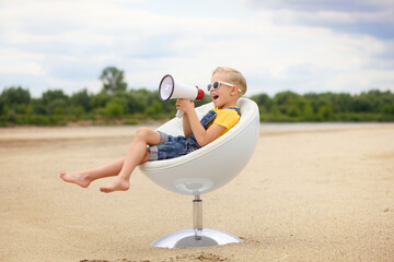 6 letni chłopiec siedzi bokiem w białym fotelu na plaży i krzyczy do megafonu.