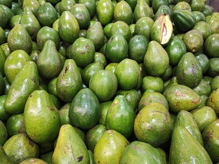 A large pile of fresh, glossy green avocados at a market. One is cut open at the top, revealing its pale yellow flesh and pit.