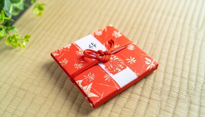 Square gift wrapped in red patterned paper with white band and red cord, resting on a woven mat with green plant.
