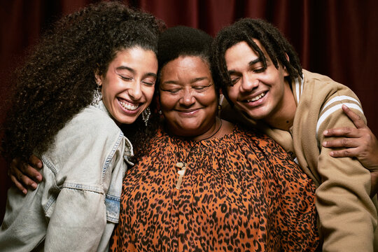 Portrait of middle aged Black woman standing between young multiethnic woman and man smiling with eyes closed in photo booth, all embracing closely in front of curtain