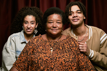 Portrait of middle aged Black woman sitting between young adult biracial woman and man smiling in photo booth, all looking directly at camera against dark curtain backdrop