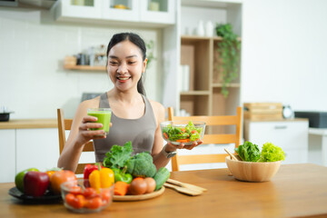 Asian woman holding green juice and apple, surrounded by fresh vegetables and salad in modern kitchen. Promotes healthy eating, fitness, and wellness lifestyle.