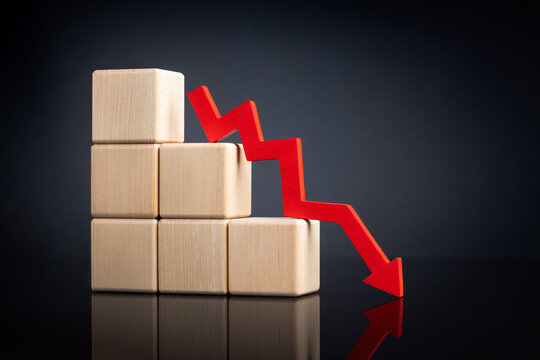 A red downward zigzag arrow next to with stacked wooden blocks, symbolizing a declining trend on a reflective dark surface. High quality photo