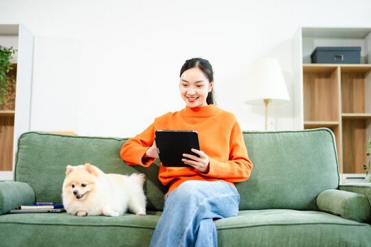 Asian woman using a tablet while relaxing on a sofa with her pomeranian dog. Cozy digital lifestyle at home with comfort, tech