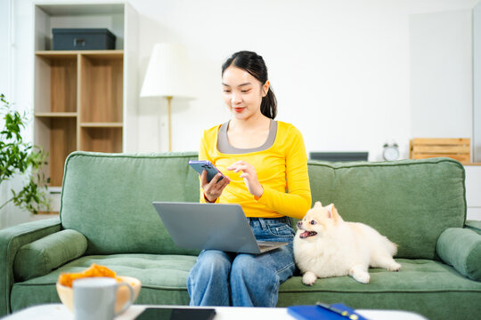 Asian woman multitasking with laptop and phone while relaxing on sofa with dog. Cozy home lifestyle with pet companionship
