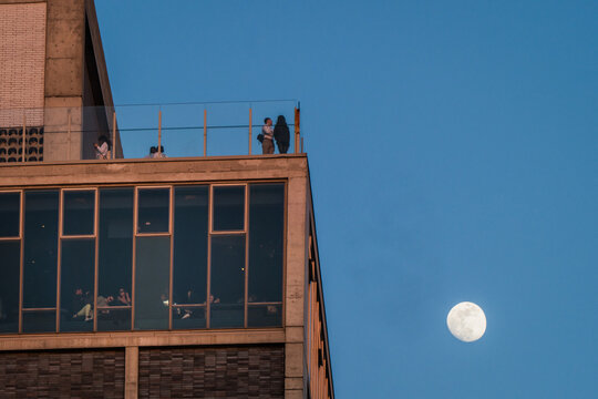 View of a modern building with a rooftop gathering and a full moon casting a soft glow against the twilight sky, New York, New York, United States.