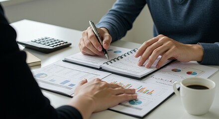 Two people reviewing financial data on a table, with a calculator, notebook, pen, and coffee.