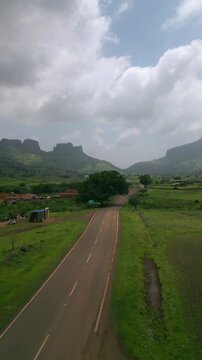 A forward-moving drone view follows a bike riding along the Nashik-Trimbak highway in Maharashtra, India. Surrounded by lush greenery and mountains, the scene unfolds under a dramatic cloudy sky