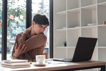 Young man feeling shoulder pain while working at modern office desk