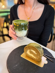 A woman holding a glass of iced matcha latte served with a slice of creamy matcha roll cake on a slate plate, creating a cozy café dessert moment.