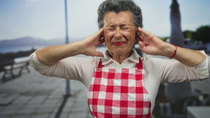 Fototapeta premium Senior woman with grey hair in apron covering ears on a seaside promenade with a worried expression, surrounded by the beach and ocean view during the day.