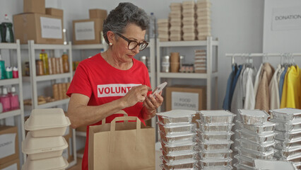 Woman preparing donations in room wearing red volunteer shirt while using mobile phone surrounded by food packages and clothes on racks highlighting community support effort.