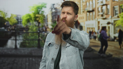 Man winking and pointing finger toward camera on city street canal walkway wearing denim jacket and beard; confidence trust boldness assertiveness.