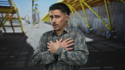 Young hispanic man outdoors city street looking scared with hands on chest under bright sunlight...