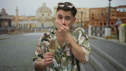 Young hispanic man in floral shirt holding ice cream, surprised in front of roman ruins on a sunny...