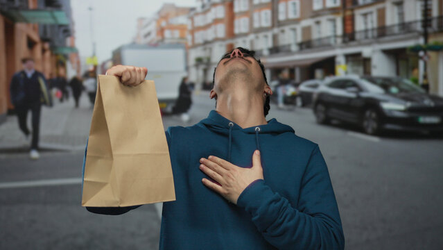 Young man celebrating with a paper bag on a city street, expressing joy with a fist pump, wearing a blue hoodie outdoors. - Powered by Adobe