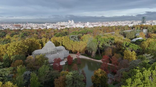 Aerial view of Palacio Cristal surrounded by autumn foliage and a serene lake, reflecting the cloudy sky, Madrid, Community of Madrid, Spain.
