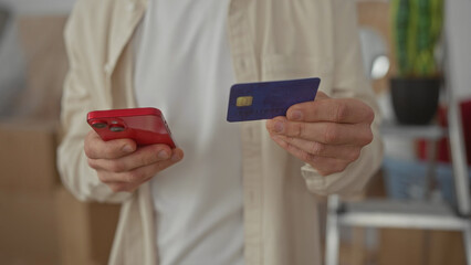 Caucasian man holding a smartphone and credit card in a new home's living room, indicating online shopping or payment activity in an indoor apartment setting.